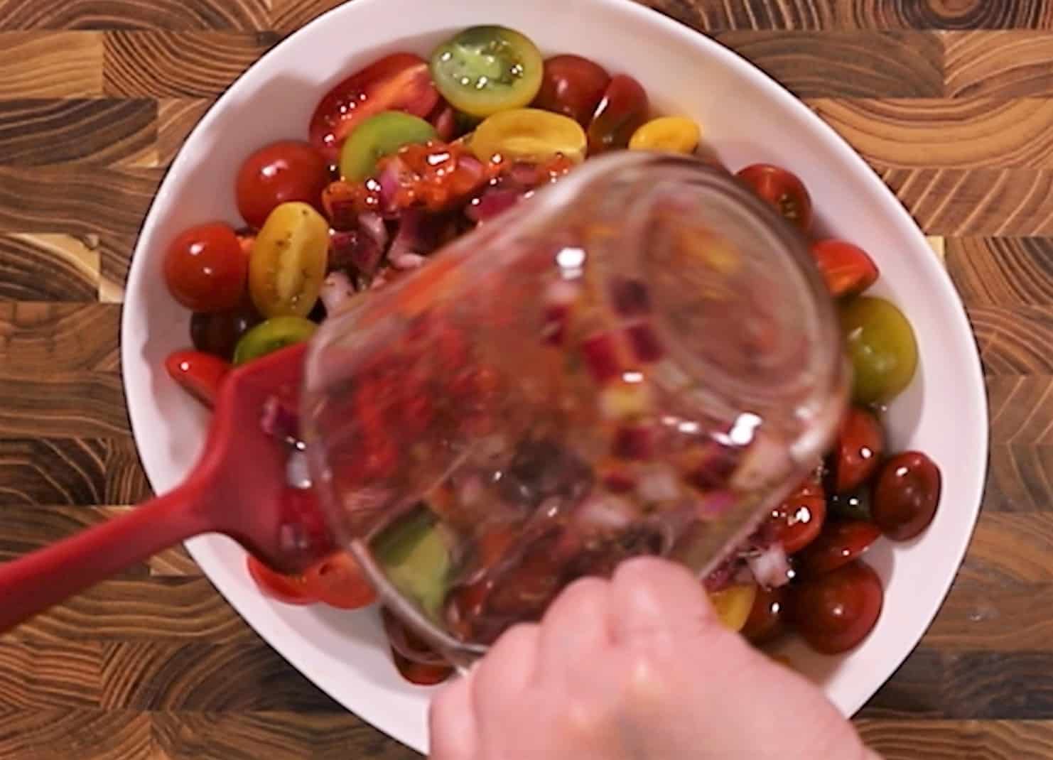 Sliced tomatoes in white serving dish with a glass measuring cup pouring marinade over the tomatoes.